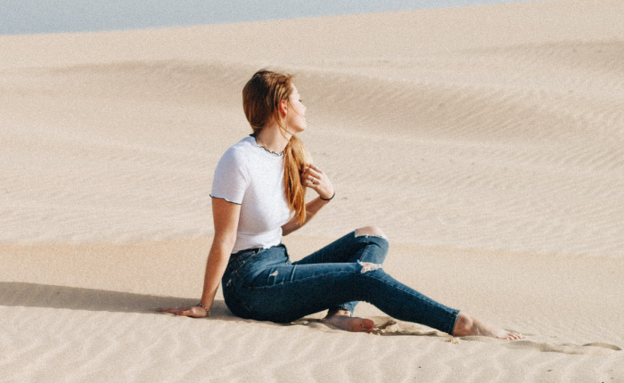 A young blonde woman looking away sits in the sand in the middle of a dry desert with a dry mouth