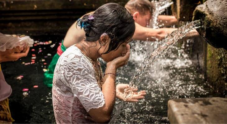 Brunette girl in a wet white shirt drinks from a stream of water flowing from a stone fountain