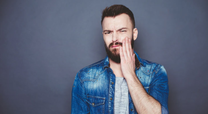 man wth a beard wearing a denim jacket holding his jaw with tooth pain